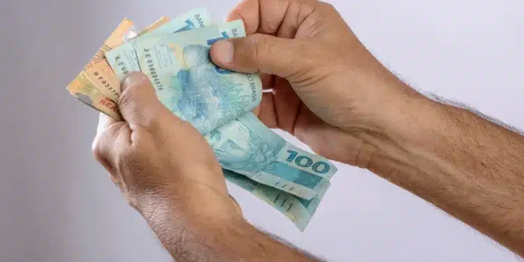 Mão segurando notas de 100 e 50 reais, representando o 13º salário e o rendimento do INSS.