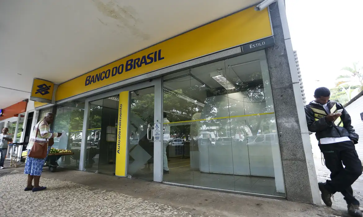 Concurso do Banco do Brasil: quando sai o edital e o que esperar 2 Agência Banco do Brasil com letreiro amarelo, portas de vidro, área de autoatendimento e pedestres na calçada