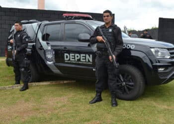 Dois policiais penais fardados e armados posam ao lado de viatura oficial do DEPEN Paraná