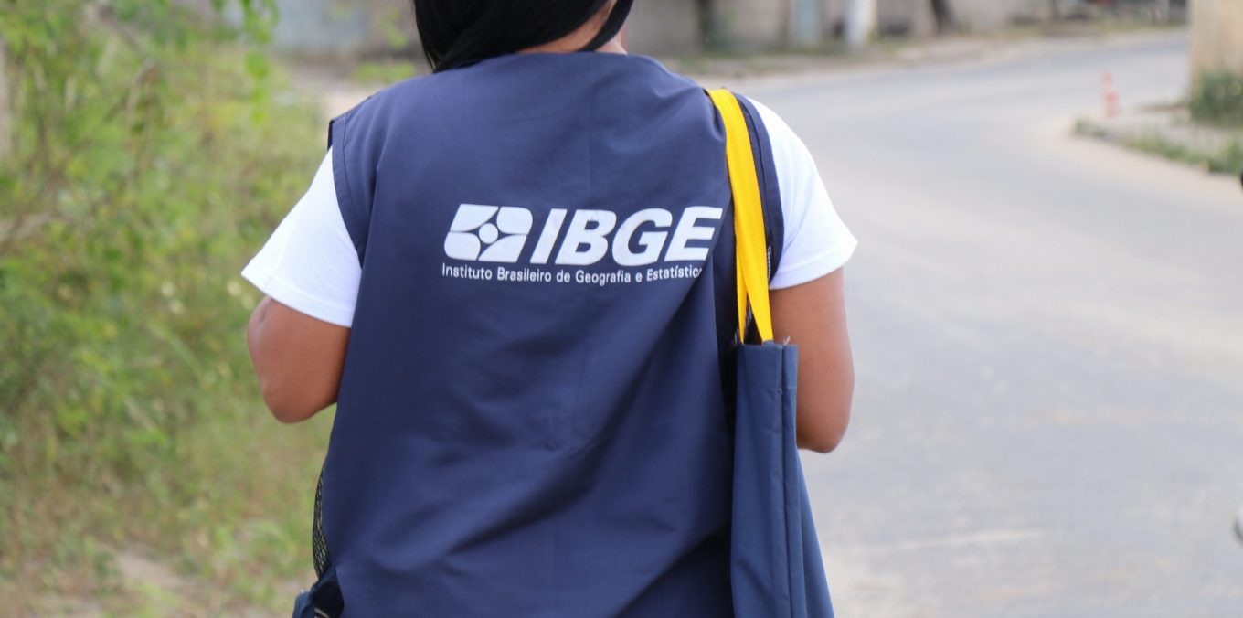 Recenseadora com colete azul do IBGE e bolsa amarela caminha por rua asfaltada em área residencial