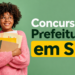 Mulher jovem de óculos e suéter rosa segura pastas e documentos com texto "Concurso Prefeitura em SP" ao fundo verde