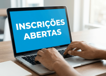 Mãos digitando em notebook prateado com tela azul exibindo "Inscrições Abertas", sobre mesa de madeira com caderno e caneca