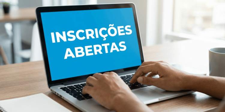 Mãos digitando em notebook prateado com tela azul exibindo "Inscrições Abertas", sobre mesa de madeira com caderno e caneca