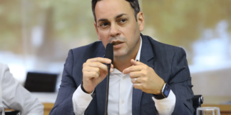 Secretário da Fazenda do Ceará de terno azul-marinho e camisa branca segura microfone durante discurso em reunião, com smartwatch no pulso