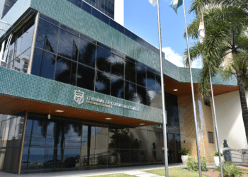 Fachada do Tribunal de Contas do RN com revestimento verde e faixas douradas, palmeiras e bandeiras