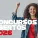 Jovem sorridente com cabelos cacheados, jaqueta jeans e mochila colorida segura um livro vermelho e ergue o punho em sinal de conquista, com a frase "Concursos Abertos 2026" em destaque, representando as oportunidades abertas nas prefeituras de Campos Novos Paulista, Bilac, Analândia e Alvinlândia