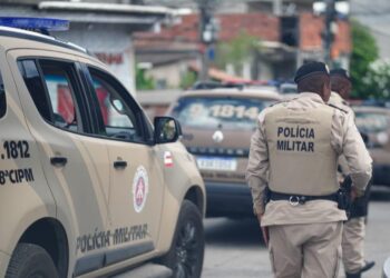 Dois policiais da PM da Bahia de costas, fardados em bege, ao lado de viatura com inscrição 8ª CIPM