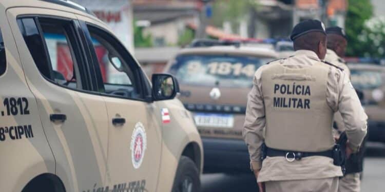 Dois policiais da PM da Bahia de costas, fardados em bege, ao lado de viatura com inscrição 8ª CIPM