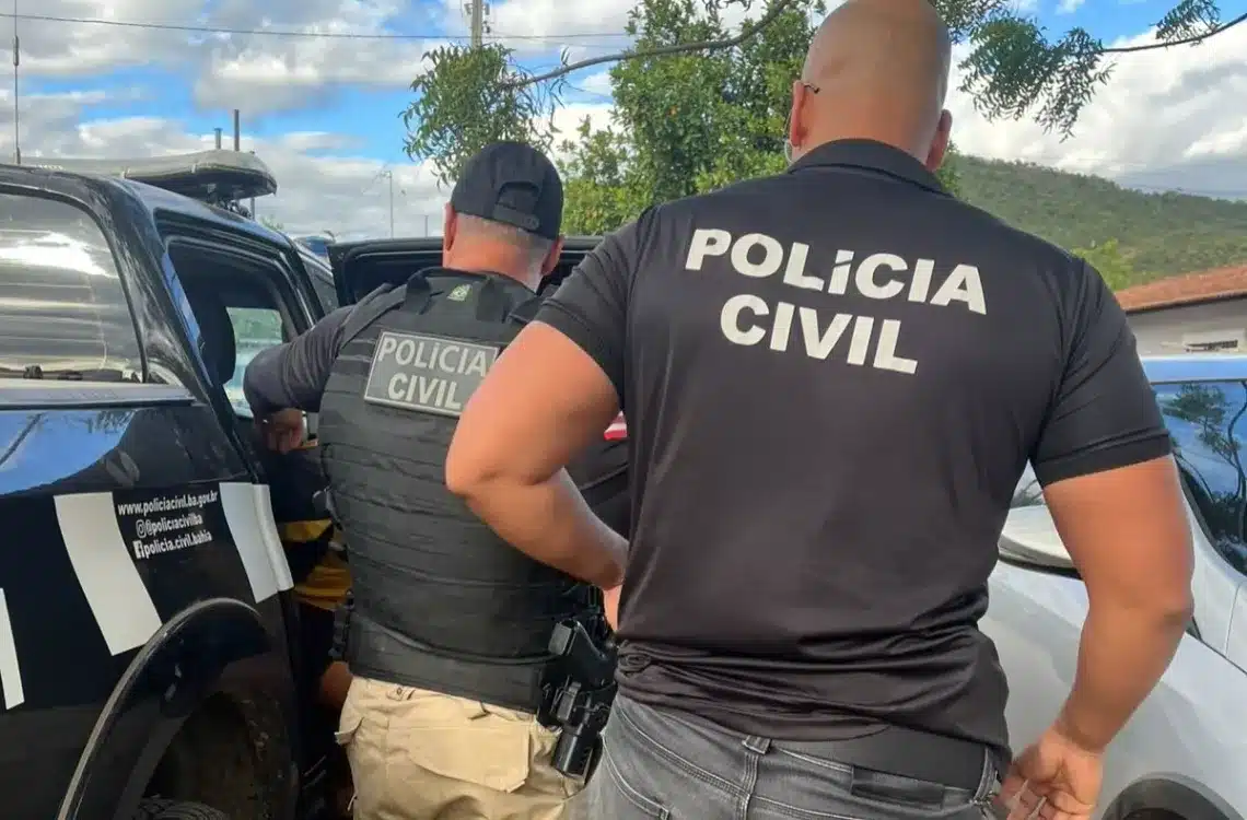 Dois agentes da Polícia Civil da Bahia de costas conduzindo suspeito até viatura preta em área externa