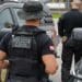 Agente da Polícia Civil de costas, com colete balístico identificado, ao lado de uma viatura durante operação policial.