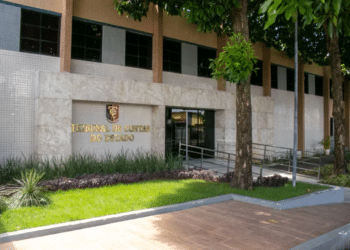 Vista externa do prédio de um Tribunal de Contas do Estado com fachada clara, árvores ao redor e placa em relevo dourado