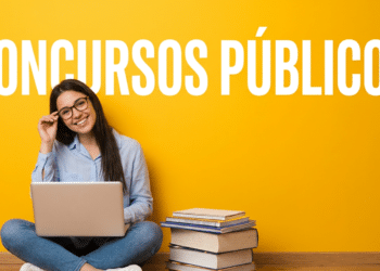 Jovem sorridente de óculos e camisa azul estuda com notebook no colo, ao lado de pilha de livros, fundo amarelo.