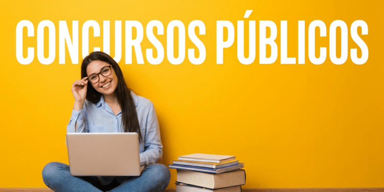 Jovem sorridente de óculos e camisa azul estuda com notebook no colo, ao lado de pilha de livros, fundo amarelo.