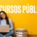 Jovem sorridente de óculos e camisa azul estuda com notebook no colo, ao lado de pilha de livros, fundo amarelo.