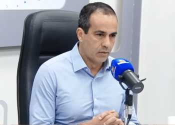 Bruno Reis, de camisa azul clara, sentado em cadeira preta com microfone azul em estúdio