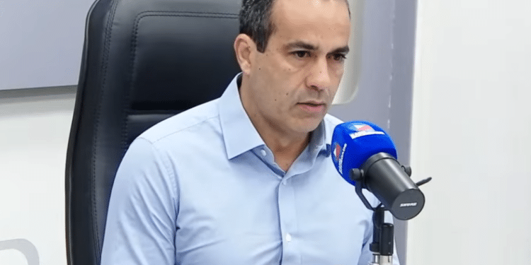 Bruno Reis, de camisa azul clara, sentado em cadeira preta com microfone azul em estúdio