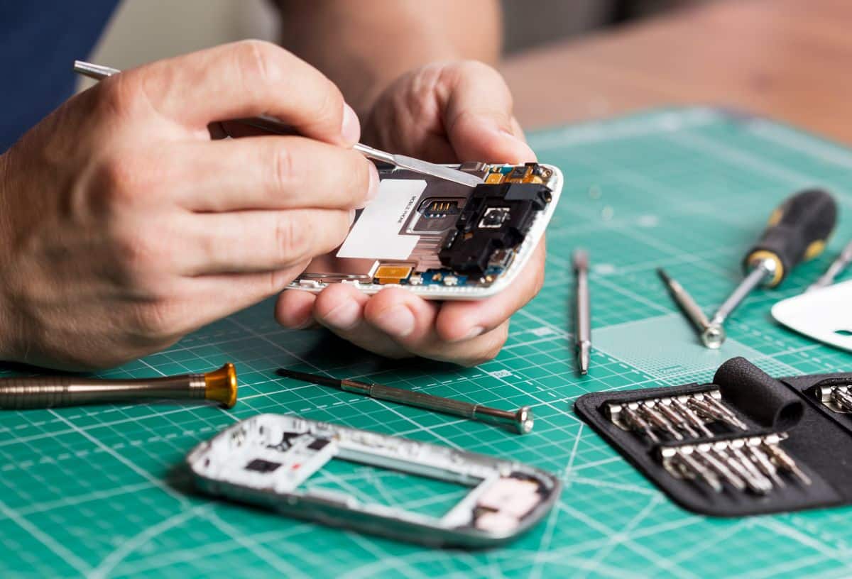 écnico consertando um celular, com foco na placa interna e ferramentas de reparo.