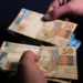 Pessoa manuseia cédulas de R$ 50, representando a primeira parcela do 13º salário do INSS que pode ser consultada diretamente pelo celular.