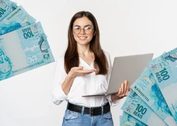 Jovem segurando notebook com notas de cem reais ao redor representando auxílio mensal de R$ 800 para cursos.