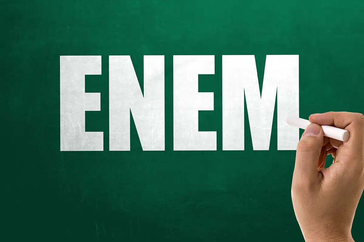 Lousa verde com a palavra ENEM destacada e uma mão escrevendo com giz, representando informações sobre o exame.