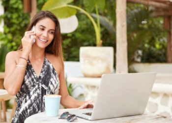 Mulher sorrindo fala ao celular enquanto usa notebook em área externa, representando rotina de trabalho flexível.