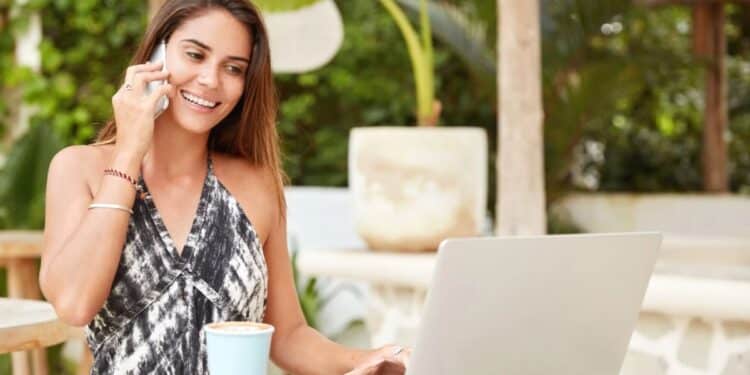 Mulher sorrindo fala ao celular enquanto usa notebook em área externa, representando rotina de trabalho flexível.