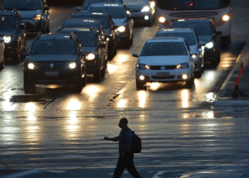 Veículos com faróis acesos parados em via molhada durante a noite, com pedestre atravessando a rua em primeiro plano