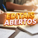 Pessoas escrevendo em cadernos sobre mesa de madeira com texto "Editais Abertos" em destaque amarelo