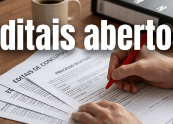 Mão segurando caneta vermelha sobre documentos de editais de concurso e processo seletivo em mesa de madeira