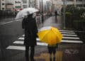 Adulto e criança com guarda-chuvas aguardam para atravessar uma rua sob forte chuva em ambiente urbano, representando os efeitos climáticos do El Niño.