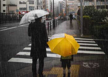 Adulto e criança com guarda-chuvas aguardam para atravessar uma rua sob forte chuva em ambiente urbano, representando os efeitos climáticos do El Niño.