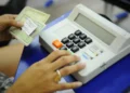 Mãos manuseiam um título de eleitor brasileiro ao lado de uma urna eletrônica em local de votação, simbolizando o exercício do voto e a regularização do documento.