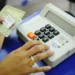 Mãos manuseiam um título de eleitor brasileiro ao lado de uma urna eletrônica em local de votação, simbolizando o exercício do voto e a regularização do documento.