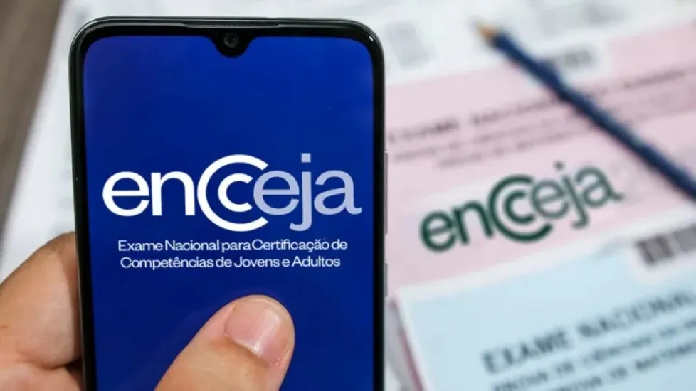 Imagem do logotipo do Encceja 2026 exibido na tela de um smartphone, com os documentos físicos do exame ao fundo.