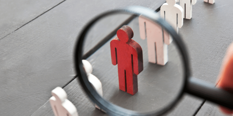 Lupa focando boneco de madeira vermelho entre peças brancas e rosas sobre mesa cinza escura