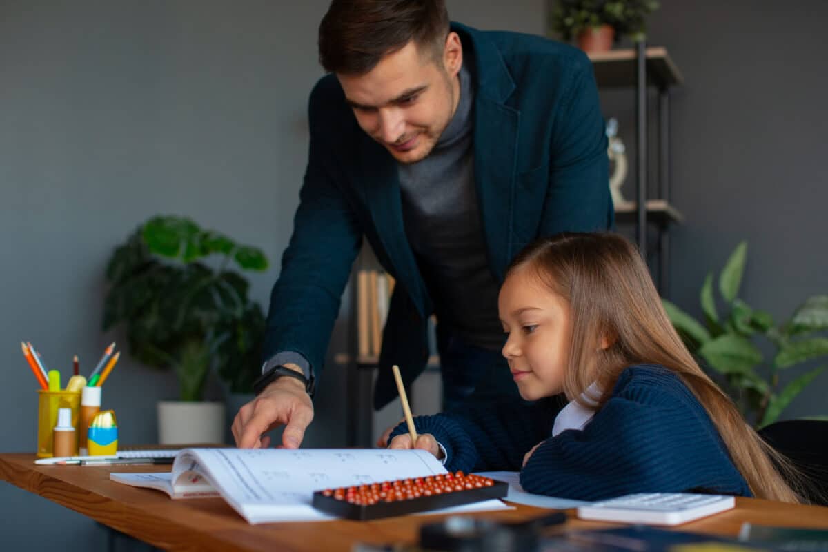 Pai orienta filha nos exercícios escolares apontando para livro aberto sobre mesa de estudos