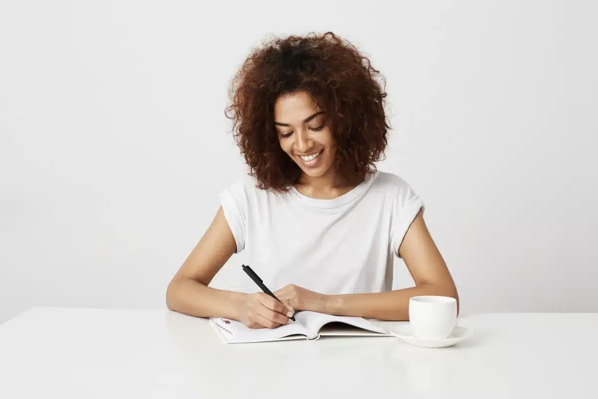 Mulher sorrindo enquanto escreve à mão em um caderno, destacando o uso de ferramentas físicas no cotidiano.