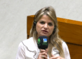 Mulher loira de cabelos longos ondulados, blusa branca com detalhes pretos, segura microfone da TV Câmara e Câmara dos Deputados