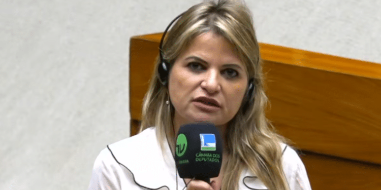Mulher loira de cabelos longos ondulados, blusa branca com detalhes pretos, segura microfone da TV Câmara e Câmara dos Deputados
