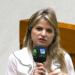 Mulher loira de cabelos longos ondulados, blusa branca com detalhes pretos, segura microfone da TV Câmara e Câmara dos Deputados