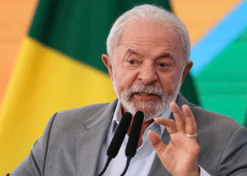 Presidente Lula gesticula durante pronunciamento com terno cinza, camisa azul e bandeiras do Brasil ao fundo
