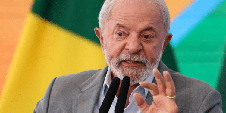 Presidente Lula gesticula durante pronunciamento com terno cinza, camisa azul e bandeiras do Brasil ao fundo