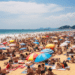 Multidão em praia de areia branca com dezenas de guarda-sóis coloridos, mar azul e arranha-céus ao fundo.