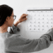 Pessoa de cabelos escuros fixando calendário mensal na parede branca