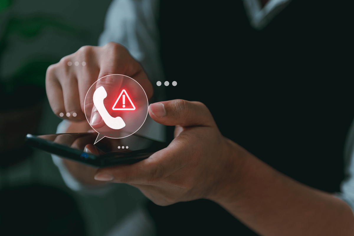 Mãos segurando smartphone preto com ícone de telefone e triângulo de alerta vermelho na tela