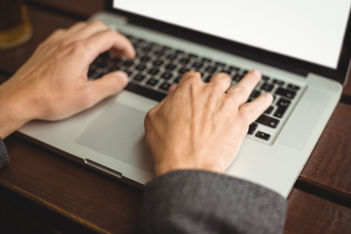 Mãos masculinas digitando em teclado de notebook prateado com tela branca, sobre mesa de madeira escura