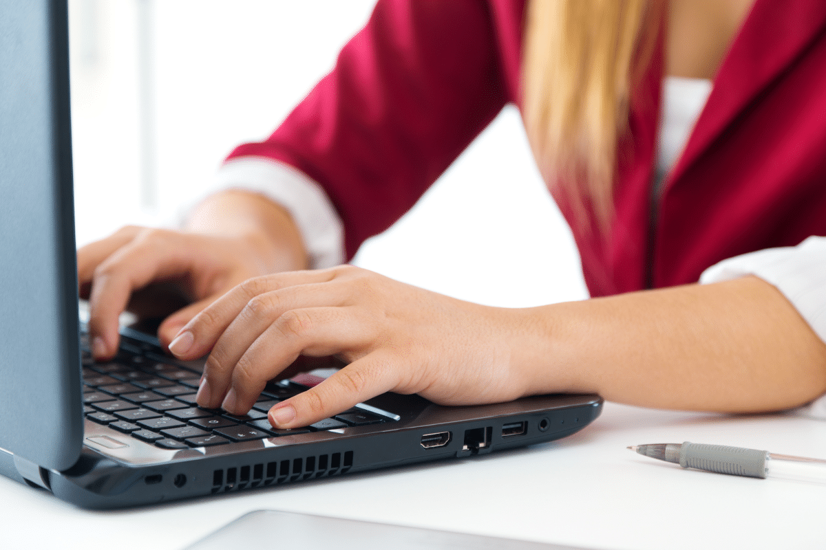 Mãos femininas digitando em teclado de notebook preto, mulher com blazer vermelho e camisa branca em ambiente de trabalho