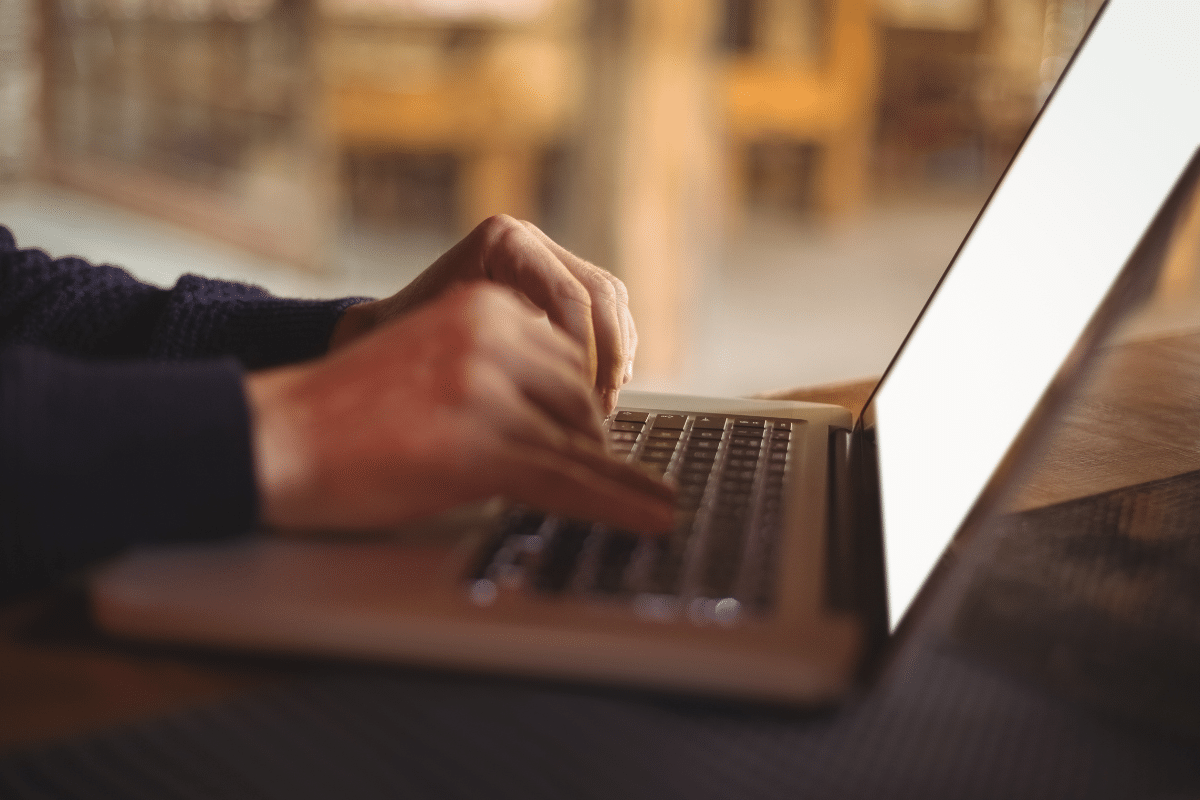 Mãos masculinas com suéter azul-marinho digitando em teclado de laptop prateado sobre mesa de madeira