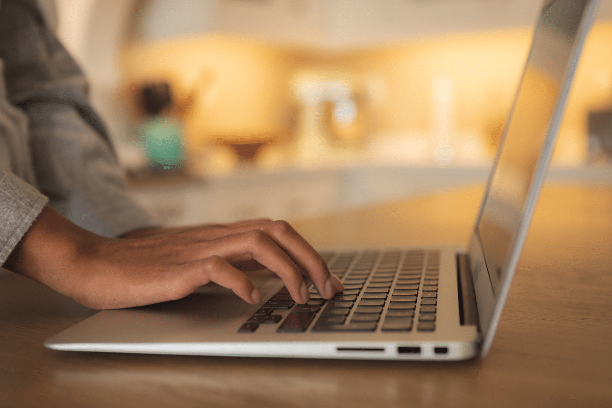 Mão feminina digitando em teclado de laptop prateado sobre mesa de madeira com iluminação quente ao fundo