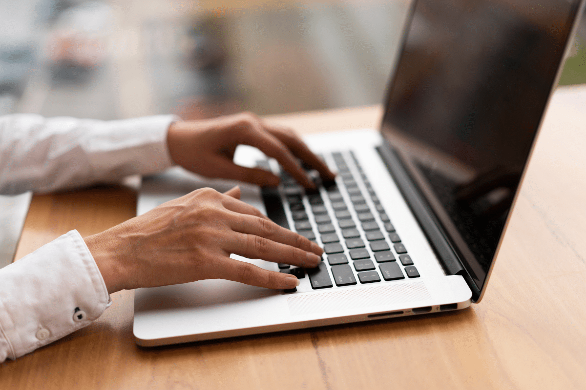 Mãos femininas com camisa branca digitando em teclado de notebook prateado sobre mesa de madeira